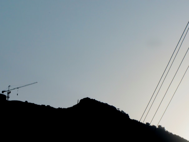A silhouette of a hillside with a crane visible on the left side, and electrical wires extending diagonally across the sky. The subtle outline of trees or vegetation is visible along the hilltop.