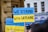 Several protest signs with blue and yellow colors expressing solidarity with Ukraine are visible. The main sign prominently displays the message 'We Stand with Ukraine,' while additional signs include text calling for visa waivers. The background features a building with arched windows.