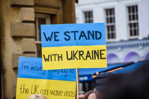 Several protest signs with blue and yellow colors expressing solidarity with Ukraine are visible. The main sign prominently displays the message 'We Stand with Ukraine,' while additional signs include text calling for visa waivers. The background features a building with arched windows.