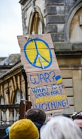 A cardboard protest sign featuring a large peace symbol painted in blue and yellow with the message 'WAR? WHAT IS IT GOOD FOR? ABSOLUTELY NOTHING!' The background includes the exterior of a historic building, and the sign is held by a person in a crowd.