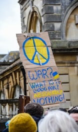 A cardboard protest sign featuring a large peace symbol painted in blue and yellow with the message 'WAR? WHAT IS IT GOOD FOR? ABSOLUTELY NOTHING!' The background includes the exterior of a historic building, and the sign is held by a person in a crowd.