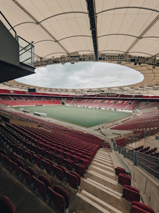 An empty football stadium with red seats and a large green field. The stands are covered by a modern, circular white canopy. The image is taken from an elevated perspective, showcasing the symmetry and structure of the arena.