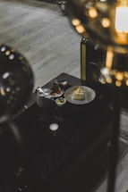 A dark, elegant setting featuring a black table with a white plate containing a pyramid-shaped dessert. Beside it are a teapot, a small cup of tea, and a small bowl with a golden element. The scene is dimly lit, creating a cozy atmosphere with blurred lights in the foreground.