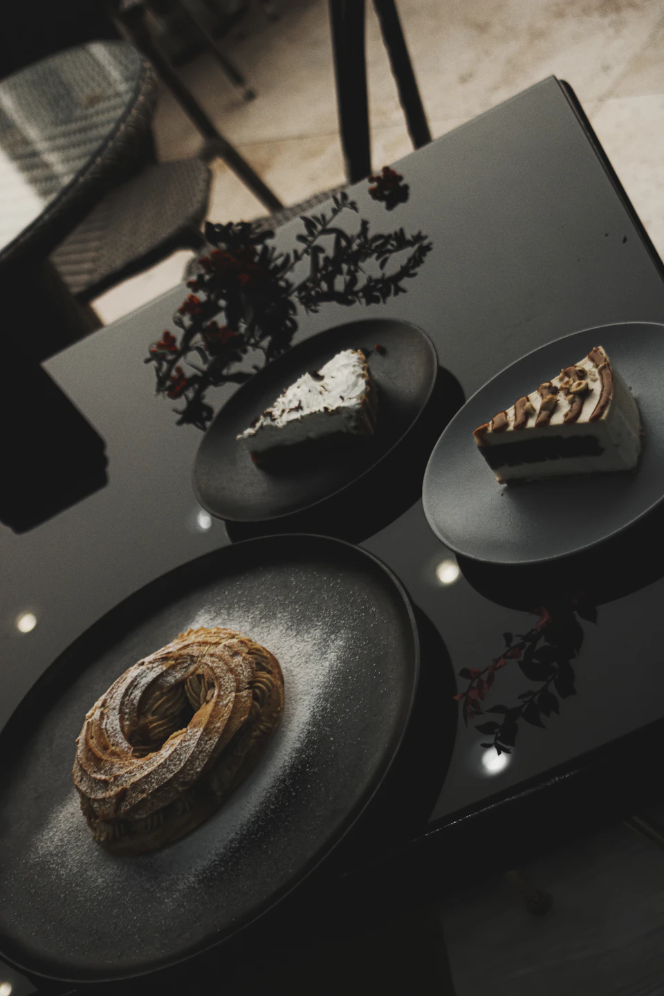 The image shows a black table with three plates of desserts arranged on it, including two slices of cake and one pastry. The cakes are covered in powdered sugar, adding to their visual appeal. One plate is positioned slightly off-center to the left, while the other two are placed symmetrically at the center of the table. In the background, there's a chair visible behind the table, suggesting that this setup might be taking place in a dining area or kitchen.
