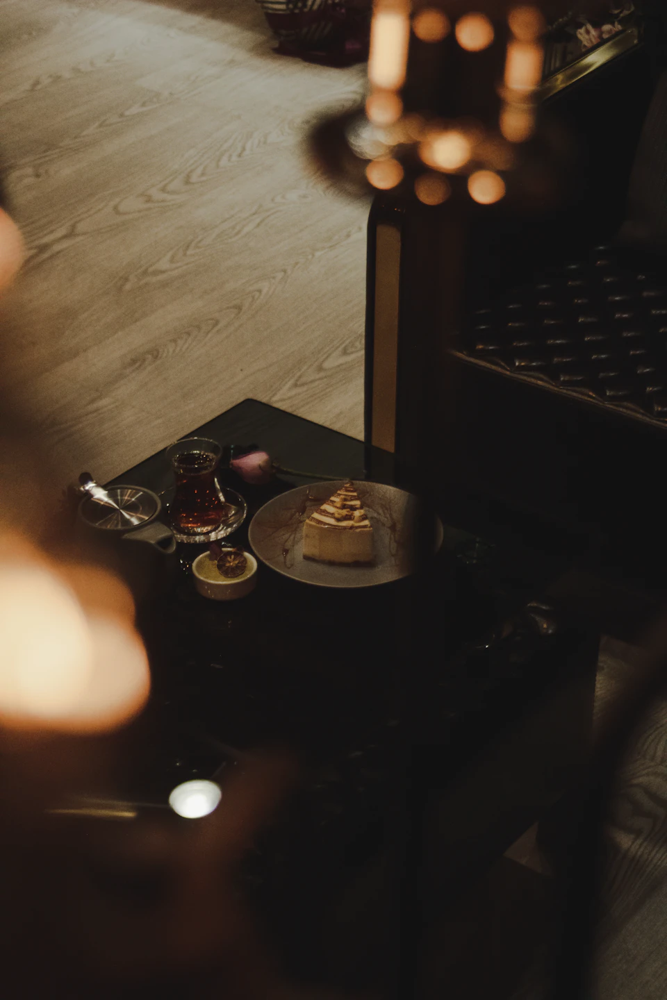 The image shows a black coffee table with a slice of cake on top, placed on a wooden floor. The cake is positioned towards the center of the table and appears to be a piece of cheesecake. To the left of the cake, there are two small bowls - one containing what looks like whipped cream or ice cream, while the other contains a spoon. A candle can also be seen in the background on the right side of the image.