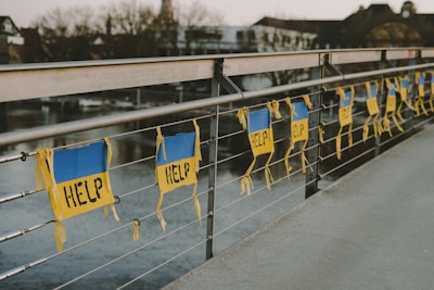 A row of signs with the word 'HELP' written on them is attached to a metal railing of a bridge. The signs have a blue and yellow color scheme and hang parallel to the railing. The background shows a river and some buildings that appear out of focus.