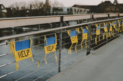 A row of signs with the word 'HELP' written on them is attached to a metal railing of a bridge. The signs have a blue and yellow color scheme and hang parallel to the railing. The background shows a river and some buildings that appear out of focus.