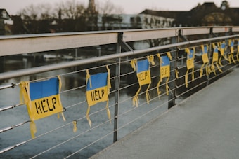 A row of signs with the word 'HELP' written on them is attached to a metal railing of a bridge. The signs have a blue and yellow color scheme and hang parallel to the railing. The background shows a river and some buildings that appear out of focus.
