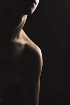 A dimly lit photograph featuring the silhouette of a person's neck and shoulder. The lighting creates a dramatic contrast between the shadowed and illuminated areas, emphasizing the contours and texture of the skin. The face is partially visible, creating a mysterious and introspective atmosphere.