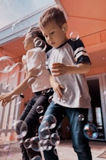 Two children are playing joyfully surrounded by bubbles. The environment appears to be cheerful and playful with bubbles of various sizes floating around them. The kids, dressed casually, seem to be having fun indoors or on a patio.