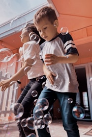 Two children are playing joyfully surrounded by bubbles. The environment appears to be cheerful and playful with bubbles of various sizes floating around them. The kids, dressed casually, seem to be having fun indoors or on a patio.