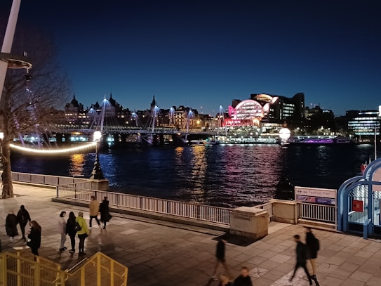 A nighttime urban scene featuring a river with illuminated bridges and buildings in the background. Vibrant lights reflect on the water, creating a picturesque cityscape. Several people are walking along a riverside path in the foreground.
