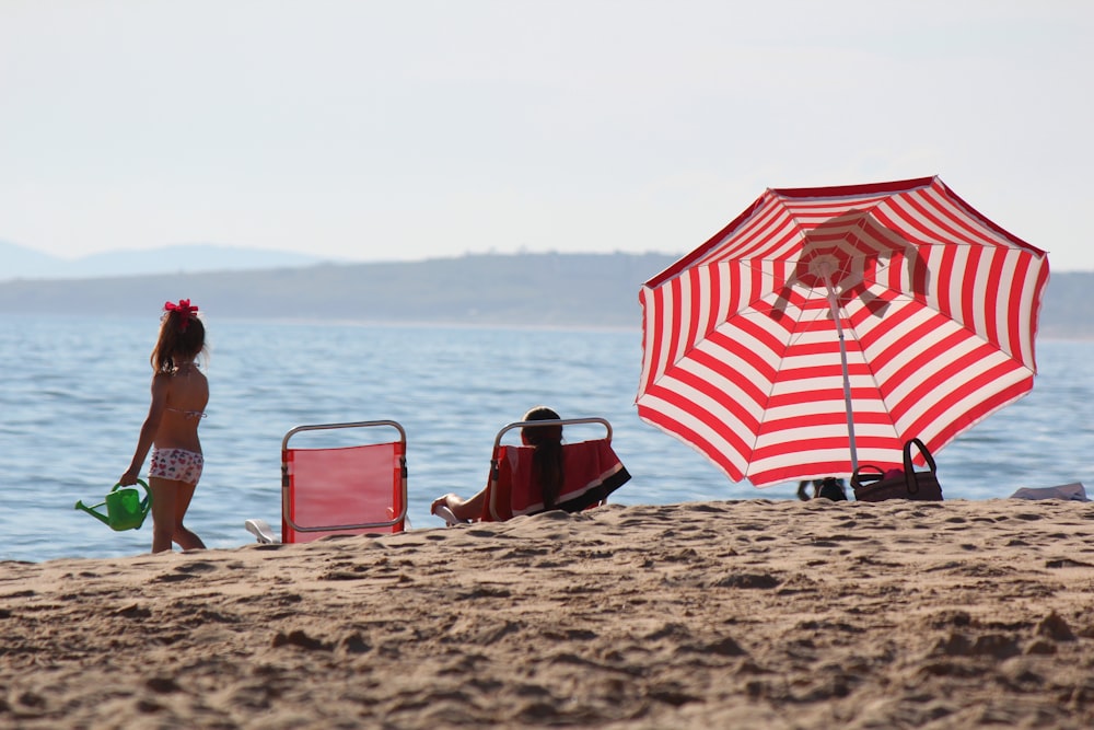 a little girl standing on a beach next to a red and white umbrella