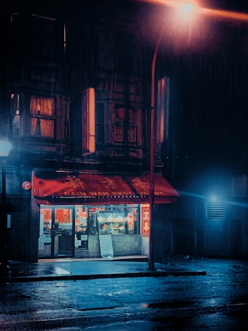 A dimly lit restaurant with a red awning and illuminated interior on a rainy night. The street is wet and reflects the light from a streetlamp nearby, adding a moody ambiance. The restaurant appears quaint with bright windows showcasing a warm interior. The overall setting is urban and somewhat nostalgic.