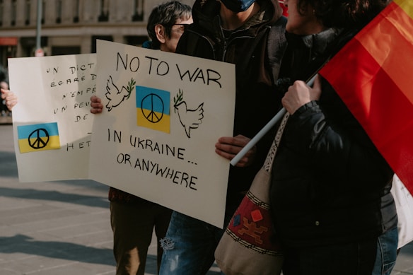 People are holding protest signs with messages advocating for peace and opposing war, specifically mentioning Ukraine. The signs feature peace symbols and illustrations of doves. A flag is partially visible, adding color to the scene.
