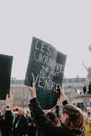 A group of people is gathered at a protest. A person is holding up a black sign with white and green writing that includes a message about Ukraine. The background features more people and some historical architecture.