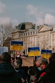 A gathering of people holding signs with messages of support. Several signs display 'I Stand With Ukraine'. Participants are wearing casual clothing and some are wearing masks. The background features a large, classic European building with a French flag on top and some leafless trees.