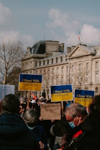 A gathering of people holding signs with messages of support. Several signs display 'I Stand With Ukraine'. Participants are wearing casual clothing and some are wearing masks. The background features a large, classic European building with a French flag on top and some leafless trees.