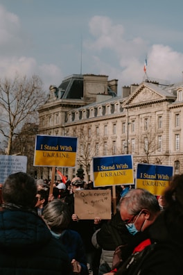 A gathering of people holding signs with messages of support. Several signs display 'I Stand With Ukraine'. Participants are wearing casual clothing and some are wearing masks. The background features a large, classic European building with a French flag on top and some leafless trees.