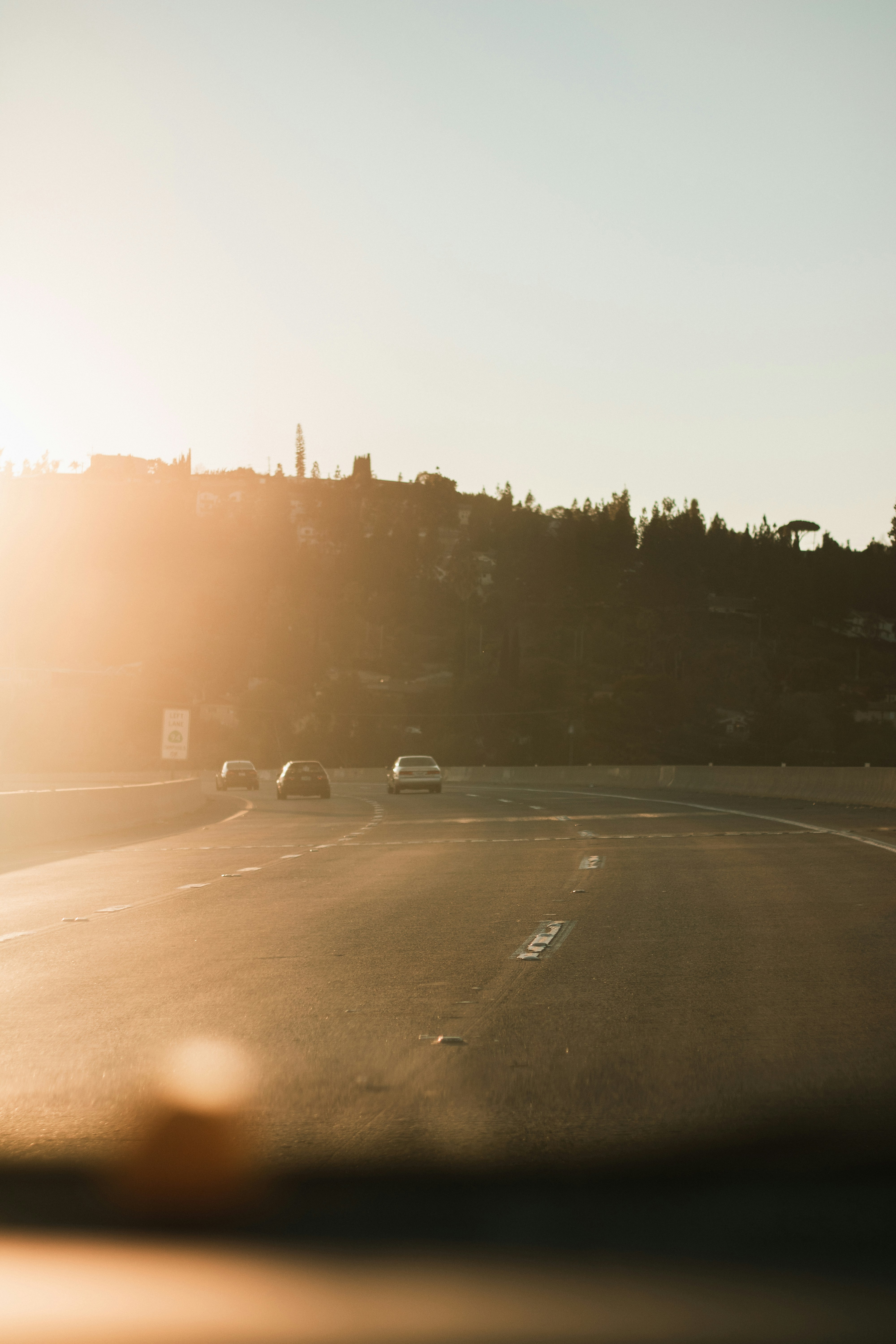 Golden-hour photograph of a sunlit highway curving toward a hillside with a few distant cars. A strong left-side lens flare washes the frame, softening foreground details.