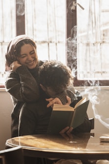A joyful scene featuring two people, one reading a book while the other affectionately embraces them from behind. Sunlight filters through a window, creating a warm and cozy atmosphere. Smoke or steam curls delicately in the air, adding a sense of tranquility and relaxation.
