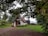 A small, quaint A-frame house is nestled among lush greenery and large trees. The house features a brick chimney, a wooden door, and a small flower garden in front. A pile of firewood is visible to the right. The grass is vibrant, and the sky is overcast, casting a serene ambiance.