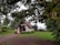 A small, quaint A-frame house is nestled among lush greenery and large trees. The house features a brick chimney, a wooden door, and a small flower garden in front. A pile of firewood is visible to the right. The grass is vibrant, and the sky is overcast, casting a serene ambiance.