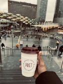 A hand holds a coffee cup from Pret, featuring maroon lettering, with an urban scene in the background. There are steps leading down to a busy square, where people are walking around. Behind the square are modern office buildings and structures with unique architectural designs.