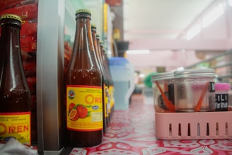 Bottles with orange beverage labels are lined up on a patterned table. A plastic basket holds jars, some with red lids and inside them are chopsticks or utensils. The setting appears to be in a casual dining area or a local eatery, with fluorescent lighting softening the ambiance.
