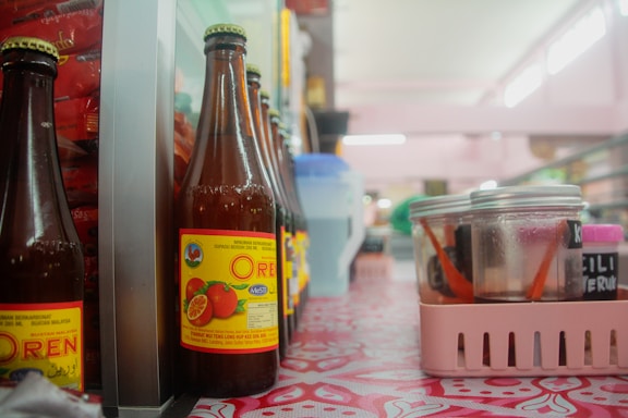 Bottles with orange beverage labels are lined up on a patterned table. A plastic basket holds jars, some with red lids and inside them are chopsticks or utensils. The setting appears to be in a casual dining area or a local eatery, with fluorescent lighting softening the ambiance.