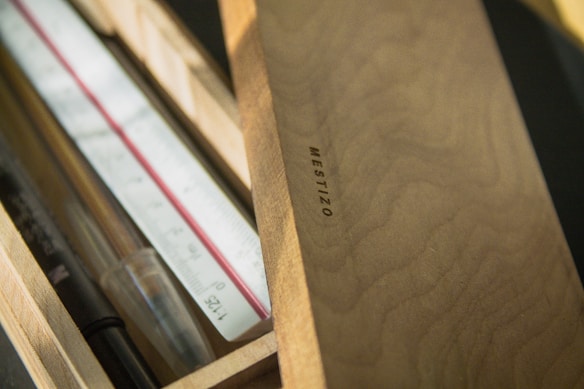 A wooden box partially open, displaying a ruler, a pen, and a clear pencil. The box is made of light wood, displaying a grain texture, and has 'MESTIZO' embossed on the lid.