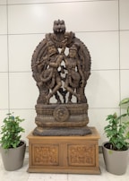 An intricately carved wooden sculpture depicting two figures in traditional attire, positioned on an ornate wooden base with detailed floral patterns. The sculpture is flanked by two potted plants, and the background features a tiled wall.