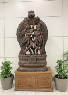 An intricately carved wooden sculpture depicting two figures in traditional attire, positioned on an ornate wooden base with detailed floral patterns. The sculpture is flanked by two potted plants, and the background features a tiled wall.