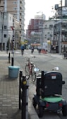 A city street scene features a mobility scooter parked on the sidewalk next to a red bicycle with a front basket. In the background, several people are cycling down the street. The area is lined with various buildings, including high-rises and smaller structures. Overhead wires crisscross the scene, and there are some traffic cones visible in the distance.
