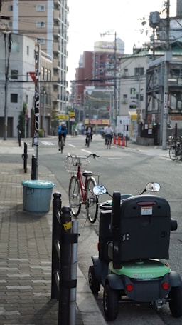 A city street scene features a mobility scooter parked on the sidewalk next to a red bicycle with a front basket. In the background, several people are cycling down the street. The area is lined with various buildings, including high-rises and smaller structures. Overhead wires crisscross the scene, and there are some traffic cones visible in the distance.