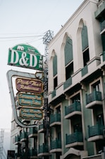 A mid-rise building with architectural features such as arched windows and green metal railings. The building has a prominent vintage-style sign featuring the name 'D&D Inn' as well as multiple attractions like a restaurant, swimming pool, beauty salon, Thai massage, and shopping place advertised. The signage is in green and gold colors, and the building appears urban with a classic design.