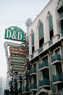 A mid-rise building with architectural features such as arched windows and green metal railings. The building has a prominent vintage-style sign featuring the name 'D&D Inn' as well as multiple attractions like a restaurant, swimming pool, beauty salon, Thai massage, and shopping place advertised. The signage is in green and gold colors, and the building appears urban with a classic design.