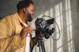 A person operating a professional camera mounted on a tripod, with sunlight casting shadows on a textured wall. The person is wearing a tan jacket and appears to be focused on their work.