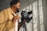 A person operating a professional camera mounted on a tripod, with sunlight casting shadows on a textured wall. The person is wearing a tan jacket and appears to be focused on their work.