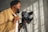A person operating a professional camera mounted on a tripod, with sunlight casting shadows on a textured wall. The person is wearing a tan jacket and appears to be focused on their work.