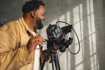 A person operating a professional camera mounted on a tripod, with sunlight casting shadows on a textured wall. The person is wearing a tan jacket and appears to be focused on their work.