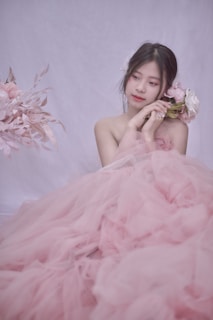 A young woman is elegantly dressed in a voluminous pink tulle gown, gently holding a bouquet of soft pink and white flowers. Her hair is styled simply, adorned with delicate floral accents, and she is gazing thoughtfully to the side. The background is a soft, light color, enhancing the romantic and serene atmosphere.