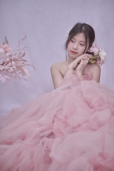 A young woman is elegantly dressed in a voluminous pink tulle gown, gently holding a bouquet of soft pink and white flowers. Her hair is styled simply, adorned with delicate floral accents, and she is gazing thoughtfully to the side. The background is a soft, light color, enhancing the romantic and serene atmosphere.