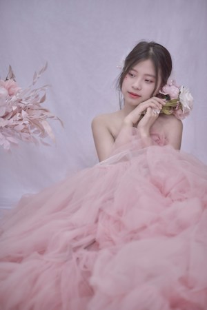 A young woman is elegantly dressed in a voluminous pink tulle gown, gently holding a bouquet of soft pink and white flowers. Her hair is styled simply, adorned with delicate floral accents, and she is gazing thoughtfully to the side. The background is a soft, light color, enhancing the romantic and serene atmosphere.