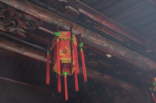 An ornate, traditional red lantern with intricate golden designs hangs from a wooden beam. The lantern is adorned with long red tassels and green decorative elements on top. The surrounding wood features carved details and a rustic, aged appearance.
