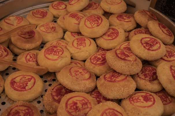 A collection of traditional round pastries with red stamps on top, likely Chinese or Asian in origin. The pastries are displayed in a large steamer basket, and some have sesame seeds on their surface. Wooden tongs are placed among the pastries, suggesting they are being served.