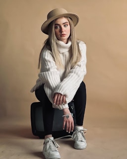 A young person with long hair wears a wide-brimmed hat and a chunky white turtleneck sweater, sitting on an old television set. The outfit is completed with black pants and white sneakers. The background is a soft beige, creating a warm and cozy atmosphere.
