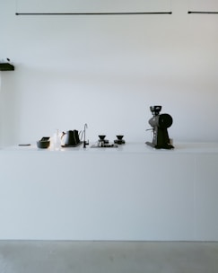 A minimalist kitchen counter with modern coffee-making equipment. On the left side, there is a small black container, a bottle of cleaner or soap, and a black electric kettle. In the center, there are two pour-over coffee drippers on a tray. On the right side, there is a large coffee grinder. The background wall is plain white, and the counter surface is also white, providing a clean and simple aesthetic.