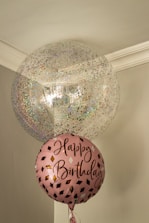 Two balloons are floating indoors, one large and transparent with multicolored confetti inside, and one smaller pink balloon with the words 'Happy Birthday' written on it in a stylish script. The setting is a corner of a room with muted colors and a simple ceiling molding.