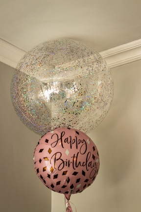 Two balloons are floating indoors, one large and transparent with multicolored confetti inside, and one smaller pink balloon with the words 'Happy Birthday' written on it in a stylish script. The setting is a corner of a room with muted colors and a simple ceiling molding.